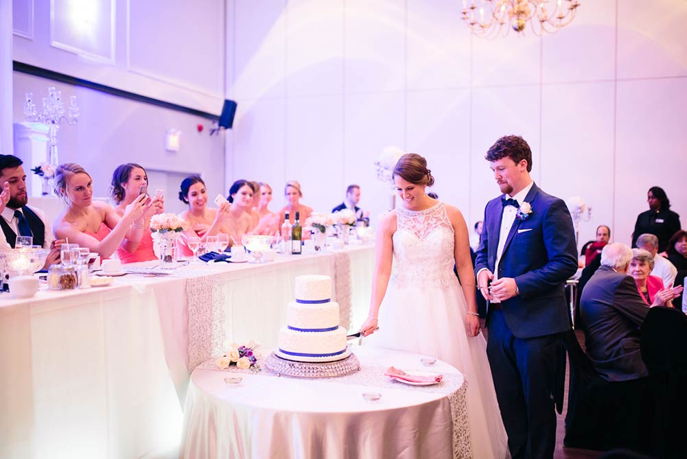 bride and groom cutting wedding cake as bridal party takes photos