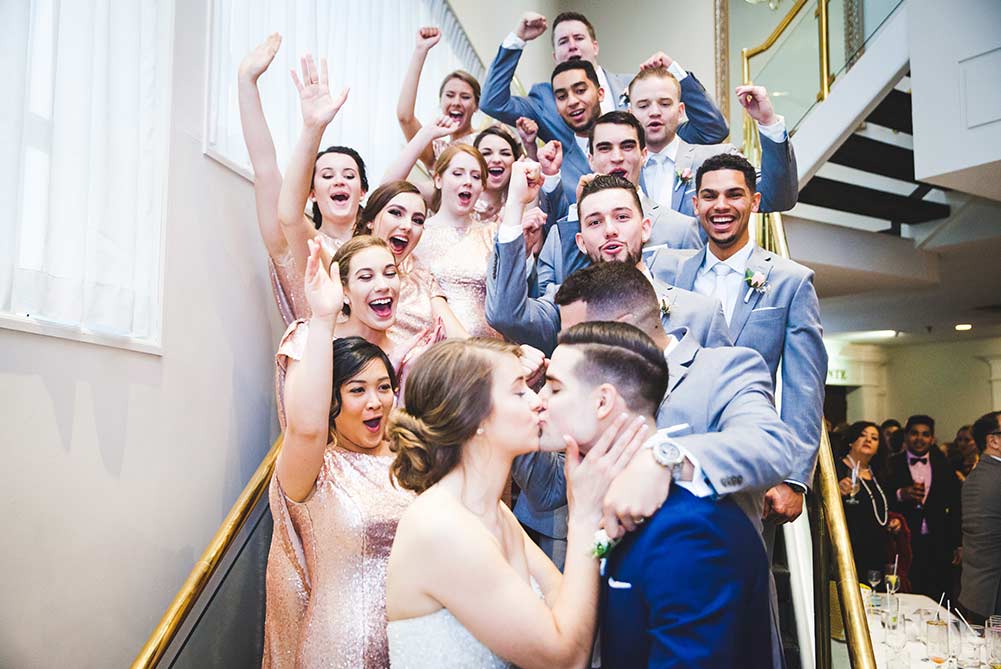 Portrait of Bride and Groom kissing on stairs as bridesmaids and groomsmen cheer
